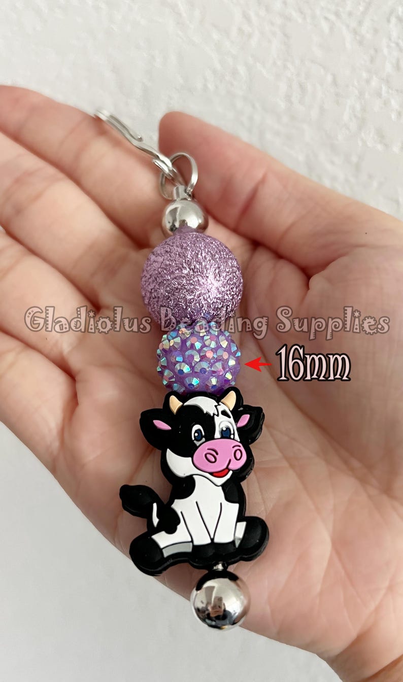 Puede incluir: Un llavero artesanal con un dije de vaca en blanco y negro, cuentas de purpurina moradas y un cierre plateado. Las cuentas miden 1,6 cm. El texto "Gladiolus Beading Supplies" es visible.