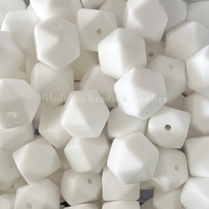 May include: A close-up shot of a pile of white, geometric beads. Each bead has a faceted, polygonal shape with a hole drilled through the center. The beads are opaque and appear to be made of a smooth material.