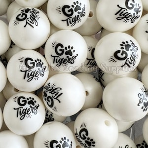 May include: A close-up of a pile of white round beads with black paw prints and the text "G Tigers" printed on them.