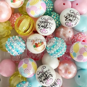 May include: A collection of colorful Easter-themed beads. The beads feature various designs, including Easter eggs, baskets, and the text "Hoppy Easter." Colors include pink, blue, yellow, and white. Some beads have a sparkly, textured surface.