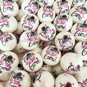 May include: A close-up of a group of white round beads with the text "Cheer for a Cure" printed in black with pink accents. The beads are decorated with a pink ribbon and a small pom-pom.