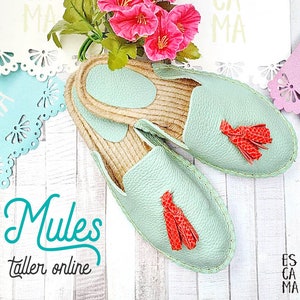 Puede incluir: Un par de mules de cuero verde claro con borlas rojas. Las mules tienen suela de yute y están sobre una superficie de madera blanca. El texto "Mules taller online" es visible en la imagen.