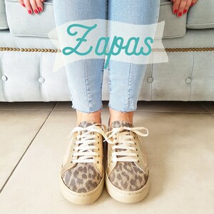 May include: A pair of gold and leopard print sneakers with white laces. The sneakers are worn by a person wearing blue jeans. The word "Zapas" is written in a white banner above the sneakers.