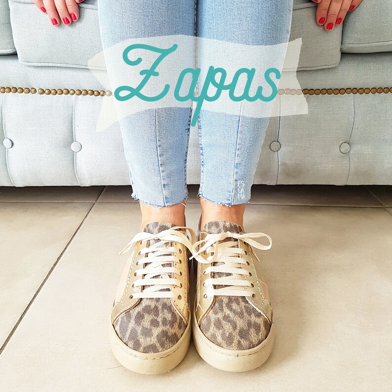 May include: A pair of gold and leopard print sneakers with white laces. The sneakers are worn by a person wearing blue jeans. The word "Zapas" is written in a white banner above the sneakers.