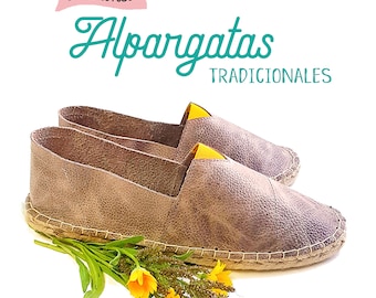 May include: A pair of brown leather espadrilles with yellow trim. The shoes are on a white background with a floral arrangement in the foreground. The text "Patrones Alpargatas TRADICIONALES" is visible above the shoes.