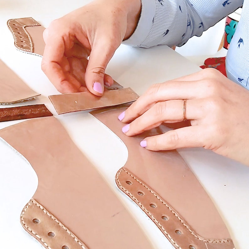 May include: A person is working on a leather craft project. They are using a pattern to cut out pieces of brown leather. The leather pieces are laid out on a white surface.
