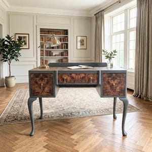May include: A desk featuring a dark grey frame and four cabriole legs, with three drawers showcasing a prominent brown burl wood veneer. The desk includes brass-toned hardware, a flat top surface, and a small back gallery rail for organisation.