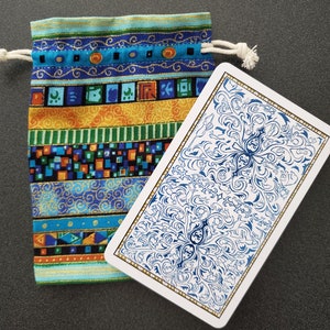 May include: A patterned drawstring pouch in blue, orange, and green, with a playing card beside it. The card has a white background with a blue floral design and gold trim. The pouch is made of fabric and has a white drawstring.