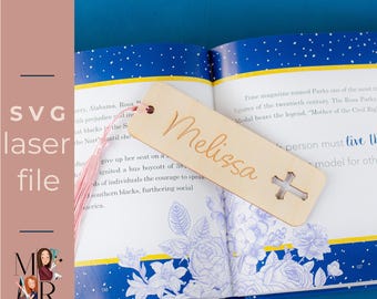 May include: A wooden bookmark with the name "Melissa" engraved on it, along with a cross-shaped cutout. The bookmark is attached to a pink tassel and rests on an open book with floral illustrations.
