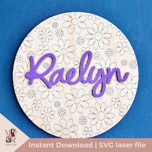 May include: A round wooden sign with a white background and a black floral pattern. The name "Raelynn" is written in purple letters on the sign.
