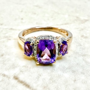May include: A rose gold ring featuring three purple amethyst gemstones. The center stone is a large, rectangular cushion cut, flanked by two smaller oval-cut stones. The ring is accented with small, clear accent stones around the center and side stones.