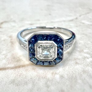 May include: A silver ring with a square-shaped diamond surrounded by a halo of blue sapphires.