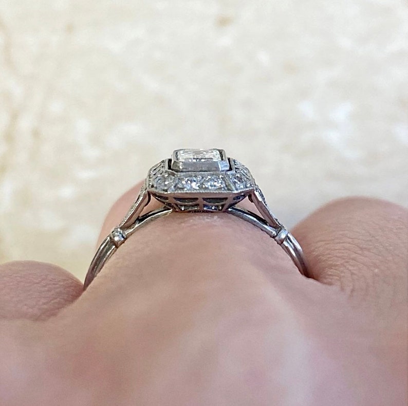 Puede incluir: Un anillo de compromiso de plata con un diamante central de forma cuadrada rodeado por un halo de diamantes m&aacute;s peque&ntilde;os. El anillo tiene una banda delicada con un detalle de milgrain.