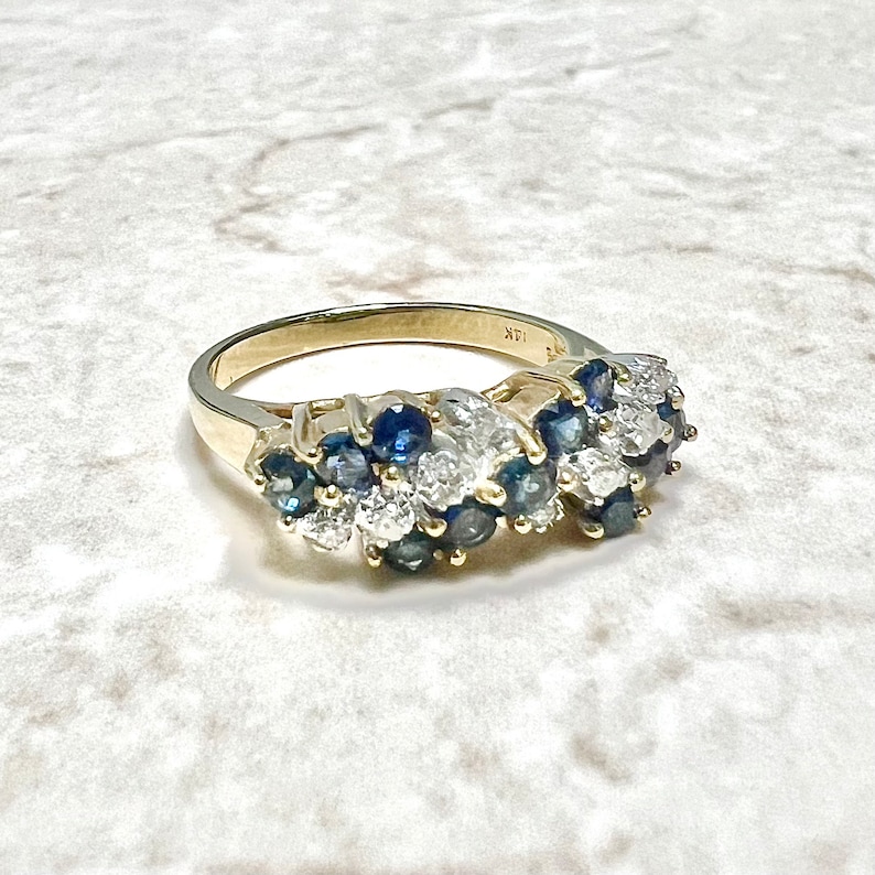 May include: A gold band ring with a cluster of blue sapphires and white diamonds. The ring is set in a pave setting and has a 14K stamp on the inside of the band.