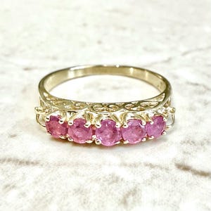 Op de afbeelding: Een gouden ring met vijf ronde, roze edelstenen op een rij. De band heeft een ingewikkeld, opengewerkt filigraanpatroon aan de zijkanten, wat een decoratief contrast vormt met de levendige, ronde roze stenen die door kleine klauwtjes worden vastgehouden.