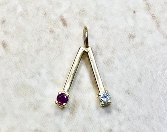 May include: A gold pendant charm shaped like a V with a red gemstone on one end and a clear gemstone on the other end.