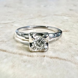 May include: A silver engagement ring with a square-shaped setting and a round, clear diamond.