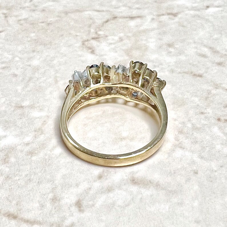 May include: A gold band ring with a row of blue and white gemstones. The ring is set with a row of round blue gemstones surrounded by smaller white gemstones.