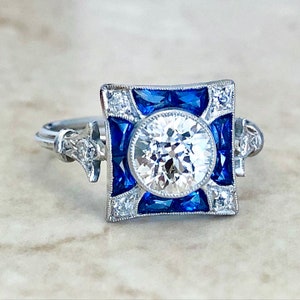 May include: A square-shaped engagement ring with a large round center diamond surrounded by a halo of smaller diamonds and blue sapphires.