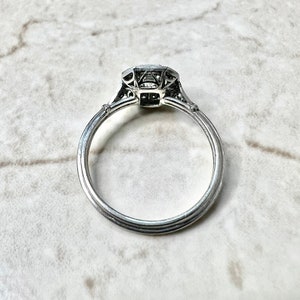 Puede incluir: Un anillo de compromiso de plata con un diamante de forma cuadrada engastado en un dise&ntilde;o delicado e intrincado. El anillo tiene una banda simple con un perfil ligeramente redondeado.
