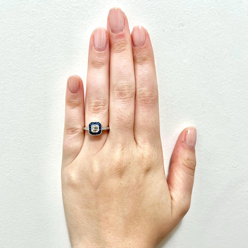 May include: A silver ring with a square-shaped center stone surrounded by a halo of smaller blue stones.