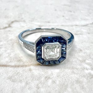 May include: A silver engagement ring with a square-shaped center diamond surrounded by a halo of blue sapphires. The band is set with smaller diamonds.