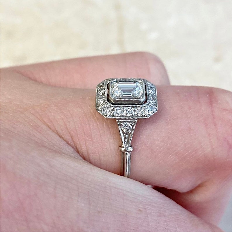 Puede incluir: Un anillo de plata con un diamante talla esmeralda de forma cuadrada rodeado por un halo de diamantes m&aacute;s peque&ntilde;os. El anillo tiene una banda delicada con un dise&ntilde;o decorativo.