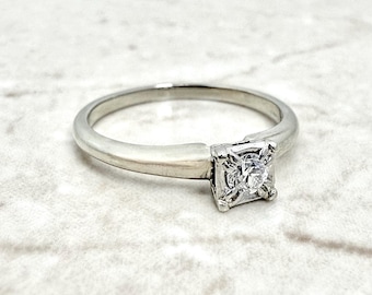 Könnte beinhalten: Ein silberner Ring mit einem quadratisch geschliffenen Diamanten. Der Diamant ist in einer quadratischen Fassung gefasst. Das Band ist schlicht und poliert. Der Ring liegt auf einer weißen Marmoroberfläche.