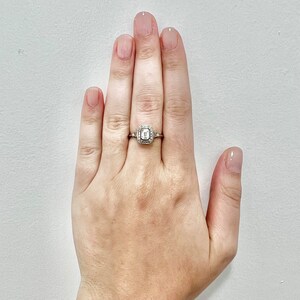 Puede incluir: Un anillo de plata con una piedra central de forma cuadrada rodeada de piedras m&aacute;s peque&ntilde;as. El anillo est&aacute; en el dedo de una persona.