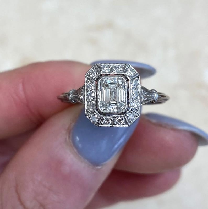 Puede incluir: Un anillo de compromiso de plata con un gran diamante talla esmeralda transparente rodeado por un halo de diamantes m&aacute;s peque&ntilde;os.