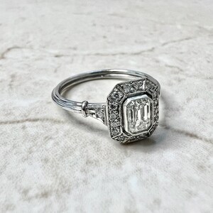 Puede incluir: Un anillo de compromiso de plata con un gran diamante rectangular de talla esmeralda engastado en un halo de diamantes m&aacute;s peque&ntilde;os.