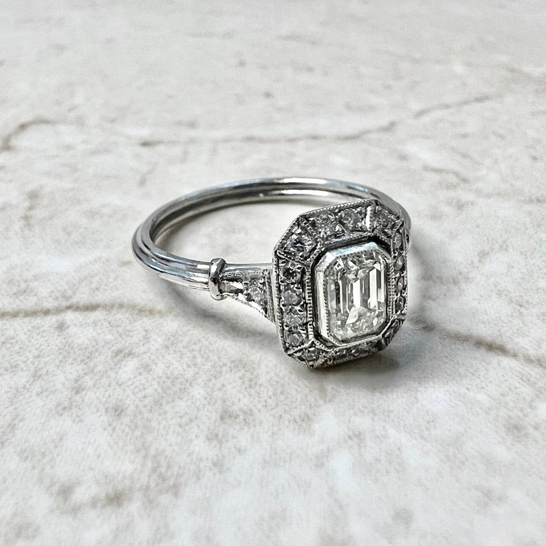 Puede incluir: Un anillo de compromiso de plata con un gran diamante rectangular de talla esmeralda engastado en un halo de diamantes m&aacute;s peque&ntilde;os.