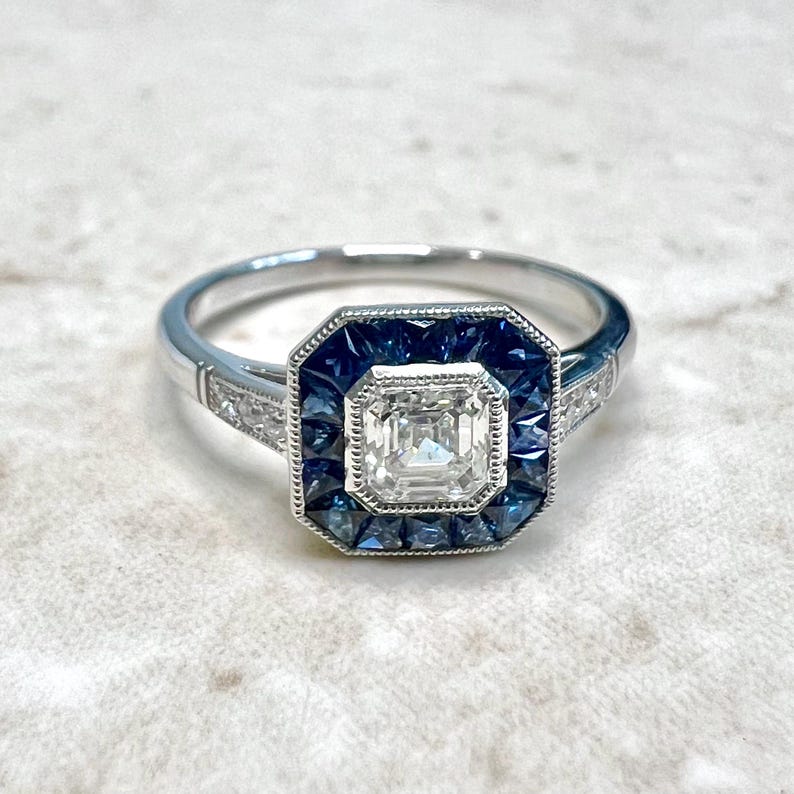 May include: A silver ring with a square-shaped center stone surrounded by a halo of blue sapphires. The ring has a delicate band with smaller diamonds.