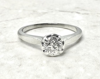 Könnte beinhalten: Ein silberner Verlobungsring mit einem runden, brillantgeschliffenen Diamanten. Der Diamant ist in einer klassischen Krappenfassung gefasst. Der Ring hat ein schlichtes, poliertes Silberband. Der Ring wird auf einer weißen, strukturierten Oberfläche gezeigt.