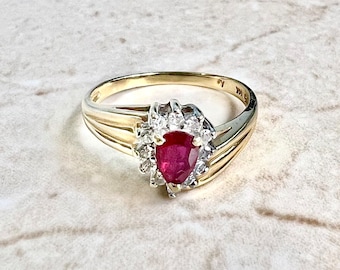 May include: A gold ring with a pear-shaped, deep red gemstone at the center, surrounded by smaller, clear gemstones. The ring's band has a ribbed design. The ring is set against a light-colored, marble-like surface.