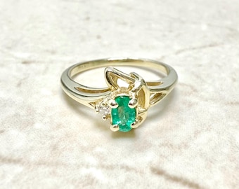 May include: A gold ring with an emerald green gemstone and two smaller diamonds. The ring has a delicate, intricate design.
