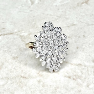 May include: A silver ring with a cluster of small, round, clear gemstones arranged in a floral or starburst pattern. The ring has a gold band and is set against a light-colored background.