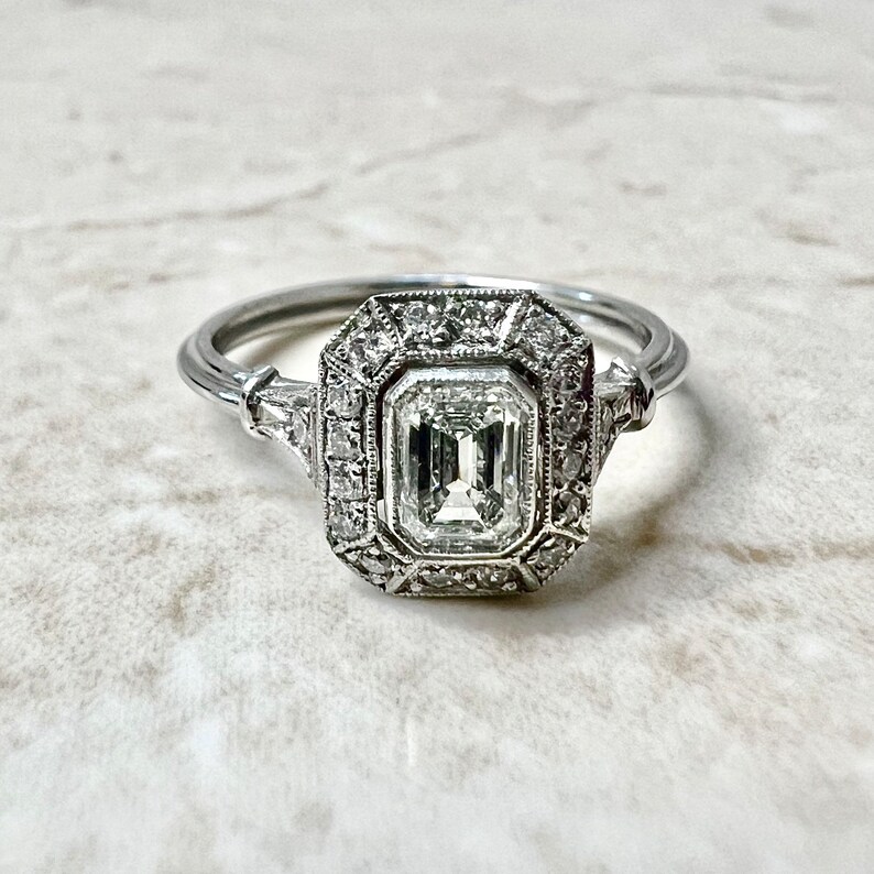 Puede incluir: Un anillo de compromiso de plata con un gran diamante talla esmeralda claro rodeado por un halo de diamantes m&aacute;s peque&ntilde;os.