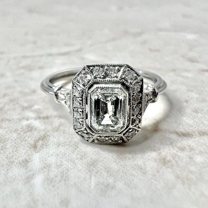 Puede incluir: Un anillo de compromiso de plata con un gran diamante talla esmeralda claro rodeado de diamantes m&aacute;s peque&ntilde;os en un engaste halo.