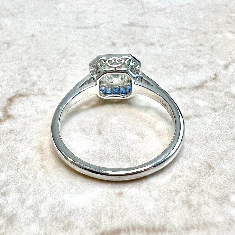 May include: A silver engagement ring with a square-shaped diamond and a halo of smaller diamonds. The band is thin and has a delicate design. The ring is set on a white background.