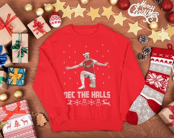 Op de afbeelding: Een rode kersttrui met de tekst "DEC THE HALLS" en een afbeelding van een persoon met een kerstmuts en een krans. De trui is omgeven door ingepakte cadeaus, versieringen en een kerstsok.