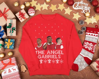 Op de afbeelding: Rode kersttrui met de tekst "The Angel Gabriels" en afbeeldingen van drie mannen. De trui is omgeven door ingepakte cadeaus, ornamenten en een kerstsok. Feestelijke vakantiekleding.