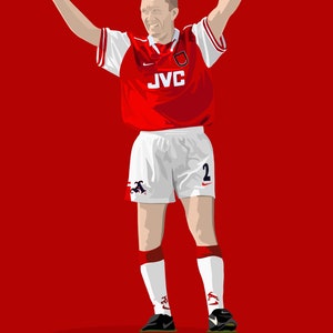 May include: A red and white soccer jersey with the text "JVC" on the front. The jersey has a white collar and white sleeves with red trim. The player is wearing white shorts with a black number "2" on the right leg. The player is raising both arms in the air.