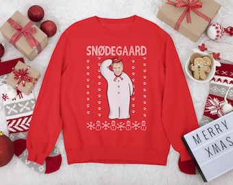 Op de afbeelding: Een rode sweatshirt met het woord "SNØDEGAARD" in witte blokletters. De sweatshirt heeft een sneeuwpop-afbeelding met het gezicht van een persoon en een rode hoed. Witte pootafdrukken en sneeuwvlokken versieren het ontwerp. Kerstartikelen omringen de sweatshirt.