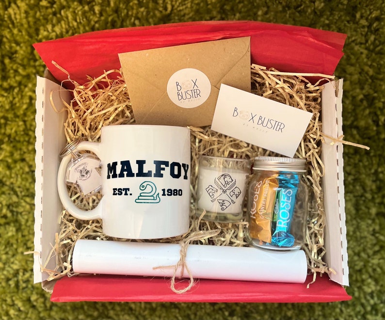 May include: A white gift box filled with brown shredded paper. The box contains a white mug with the text "MALFOY EST. 1980" and a green snake logo, a small glass candle with a black and white design, a clear glass jar with chocolate bars, and a white rolled-up paper with twine.