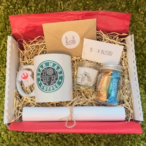 May include: A red and white gift box filled with Star Wars themed gifts. The box contains a white mug with a green Star Wars Coffee logo, a small candle with a white R2-D2 design, a glass jar with chocolate candies, and a white rolled up piece of paper.