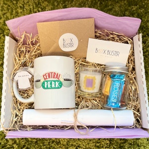 May include: A gift box filled with Friends themed items, including a mug that says "Central Perk", a candle with a purple frame, a small jar of chocolates, and a Friends keychain. The box is lined with shredded paper and has a purple tissue paper lining.