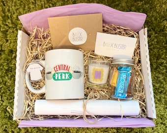 May include: A gift box filled with Friends themed items, including a mug that says "Central Perk", a candle with a purple frame, a small jar of chocolates, and a Friends keychain. The box is lined with shredded paper and has a purple tissue paper lining.