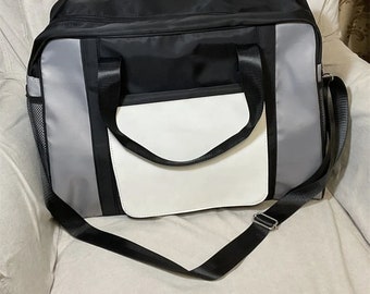 May include: A black and gray travel bag with a white leather patch on the front. The bag has a long shoulder strap and two top handles.