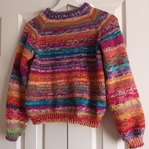 Puede incluir: Un suéter de punto a rayas de colores con cuello redondo y mangas largas. El suéter está hecho con una variedad de colores, incluyendo rojo, naranja, amarillo, verde, azul, morado y rosa.